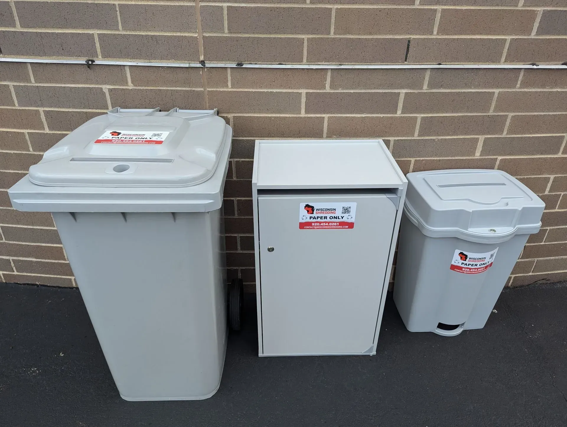 Three gray waste bins against a brick wall. The bins have lids, and stickers.