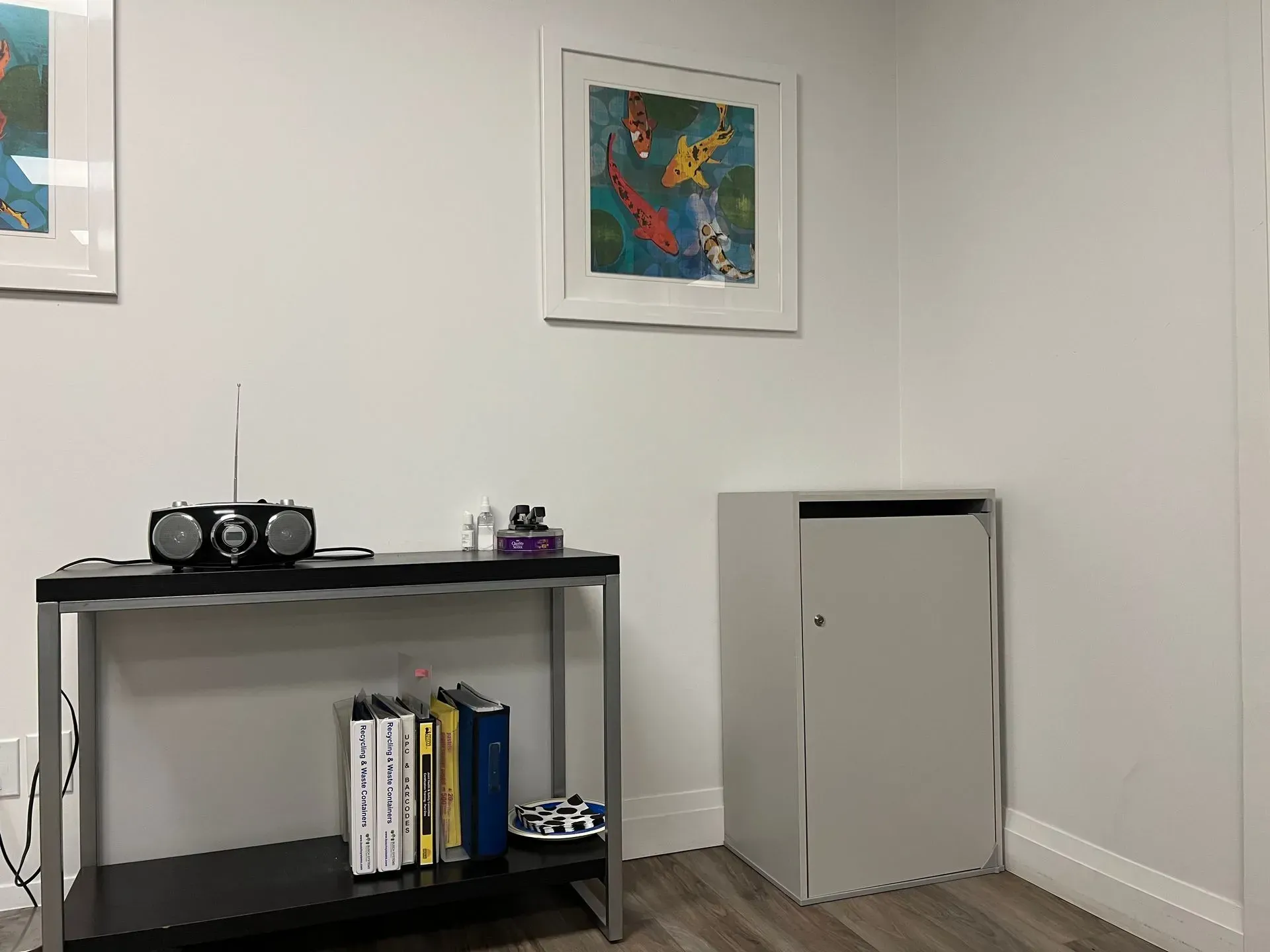 A small table with a radio and books next to a gray cabinet in a room with art on the wall.