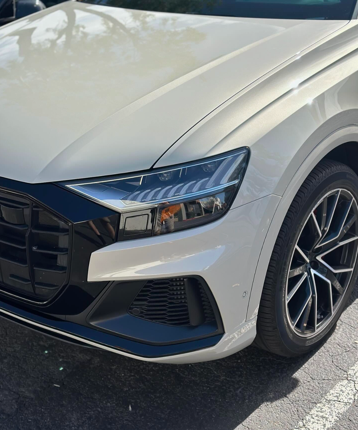 Close-up of a white sedan’s front corner, showing headlight, grille, and wheel in a parking lot.