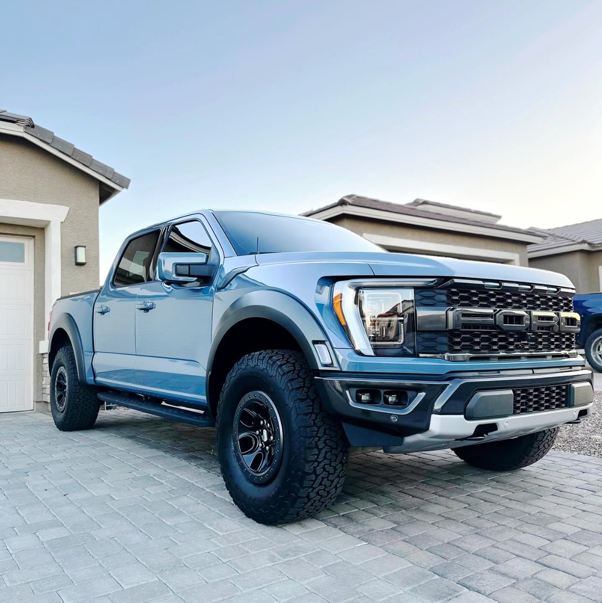 Blue Ford Raptor pickup parked in a driveway beside a house, front three-quarter view