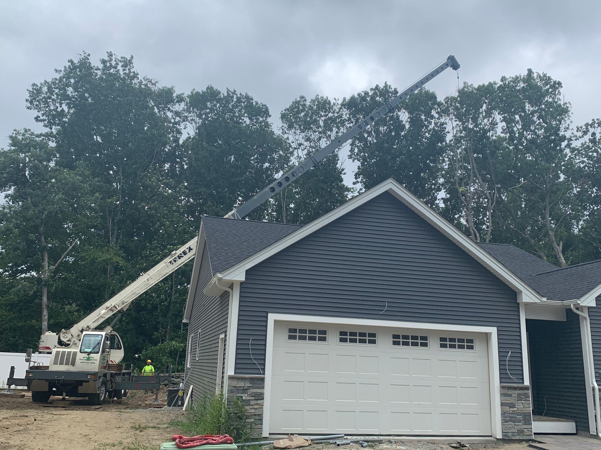 Construction site: crane lifting roofing material onto a gray garage; overcast sky.