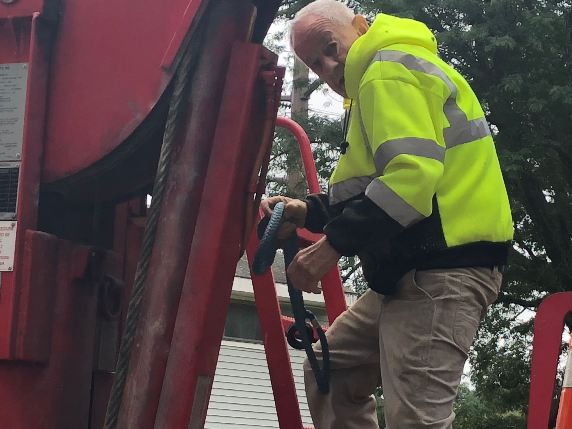 Man in neon jacket climbs garbage truck, holding a blue cord.