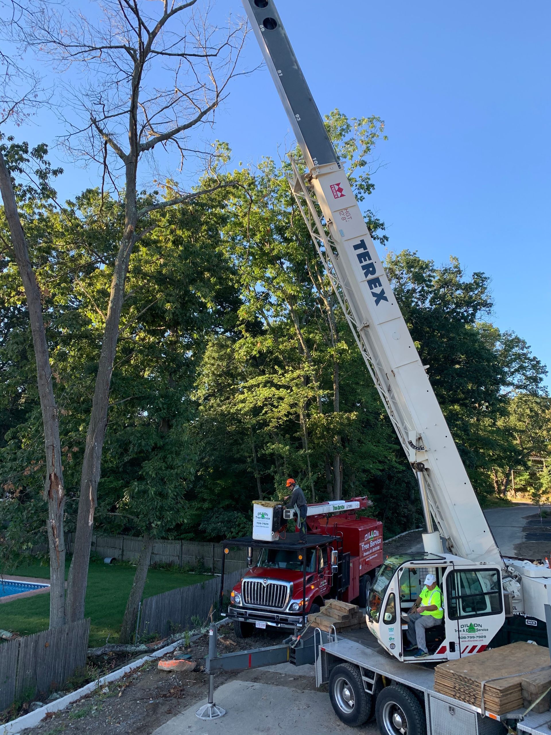 A crane lifting a red truck near a tree and woods, with a worker in the truck.