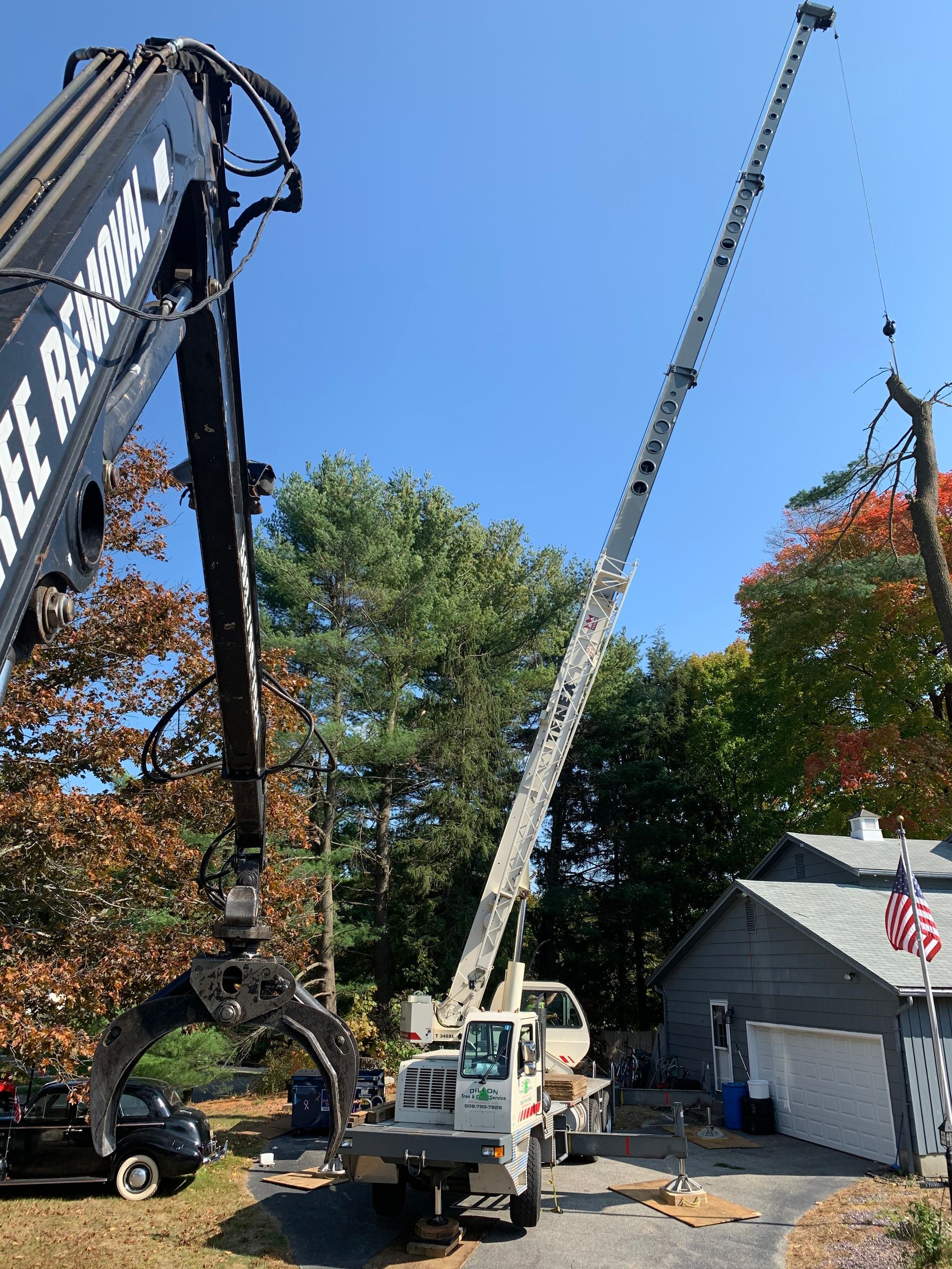 Two tree service trucks with extended booms; one has a grabber, the other has a bucket, beside a house and trees.