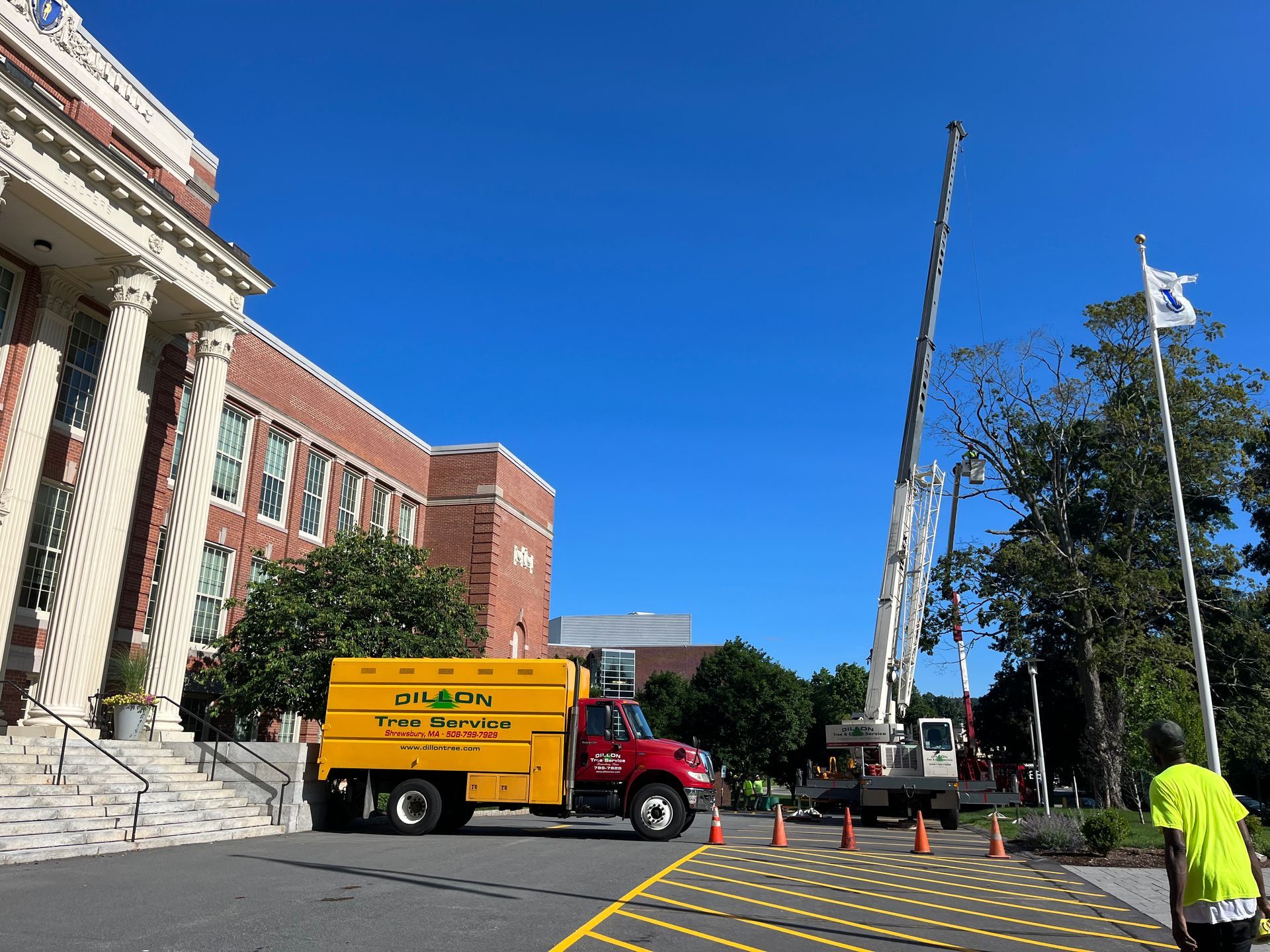 A yellow tree service truck and crane are parked near a red brick building under a blue sky.