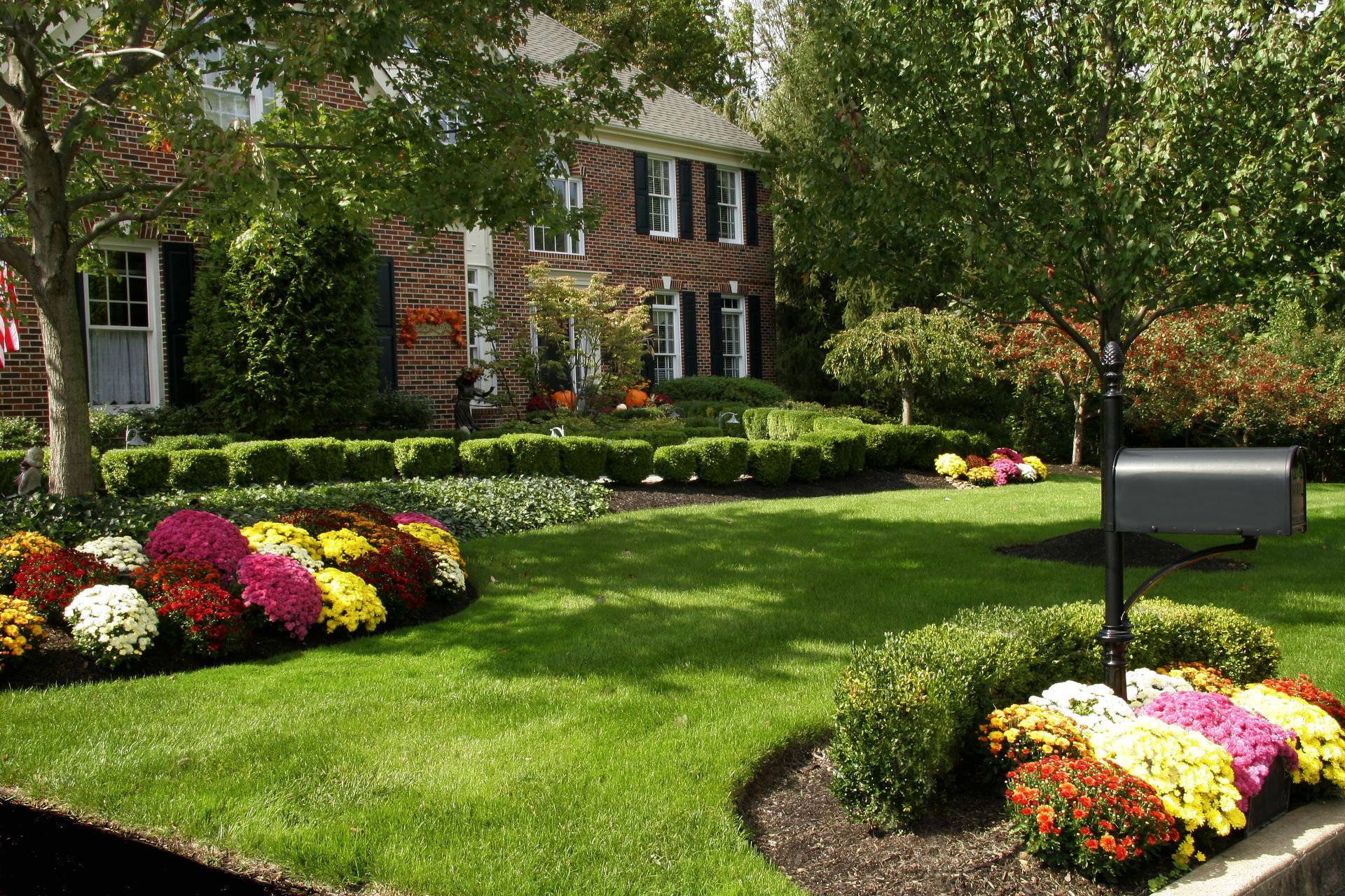 Brick house with manicured lawn, colorful flower beds, and a mailbox under leafy trees
