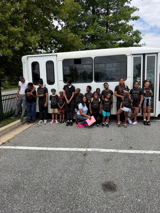 Group posing in front of a white bus on a paved roadside, with trees and a fence behind them.