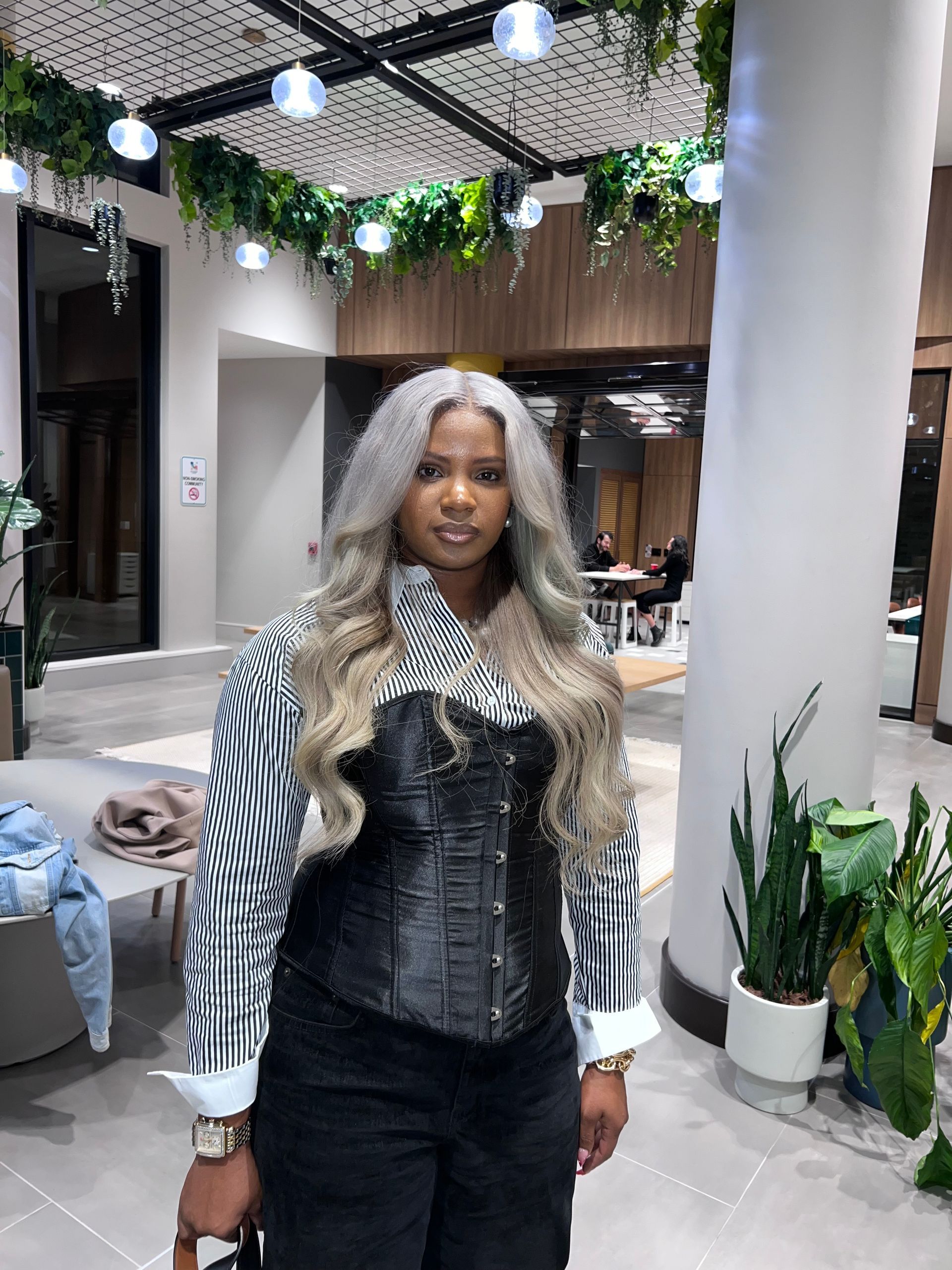 Woman in black corset and striped sleeves standing indoors with plants and hanging lights