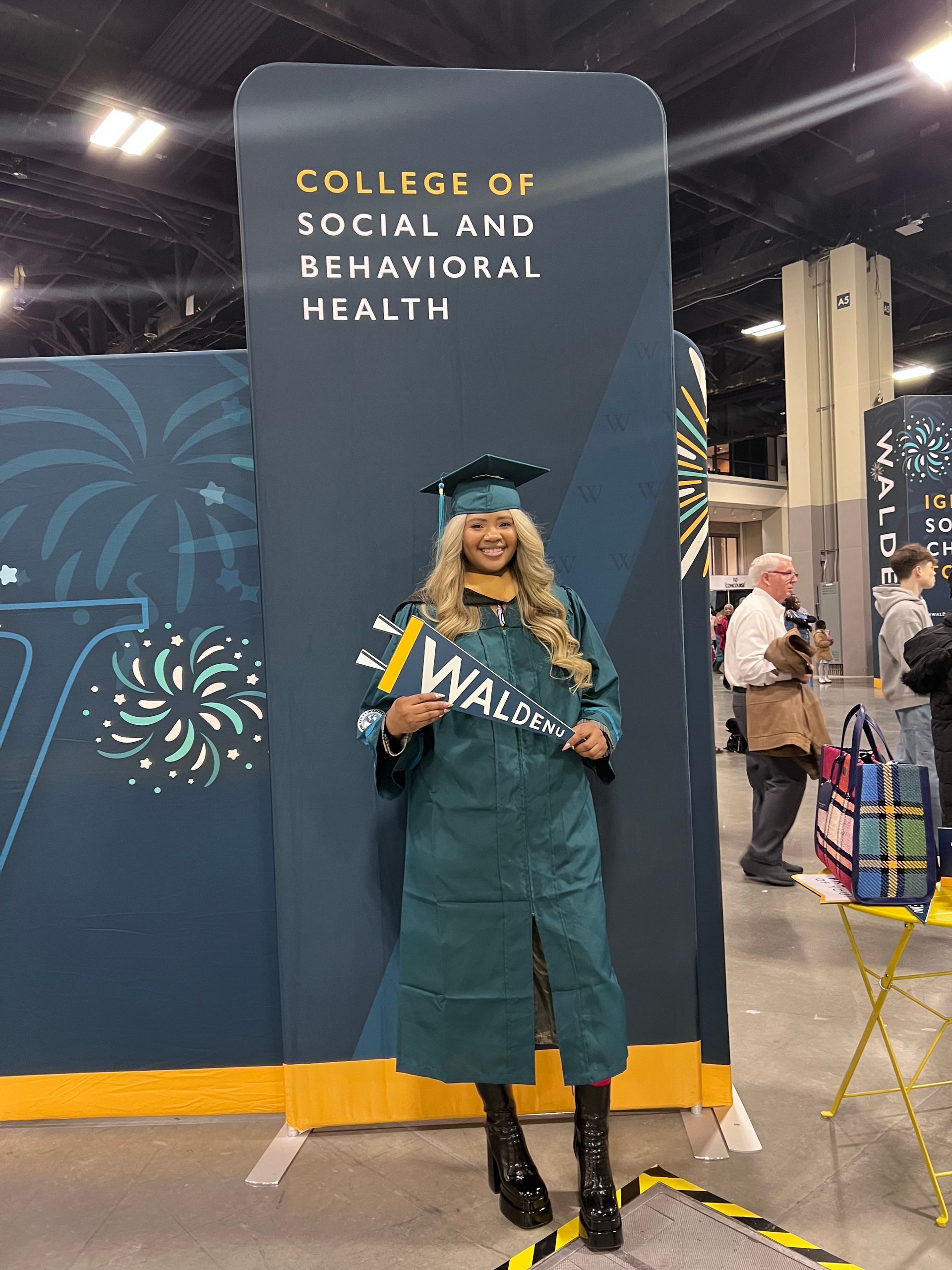 Graduate in teal cap and gown stands smiling at a college of social and behavioral health backdrop.