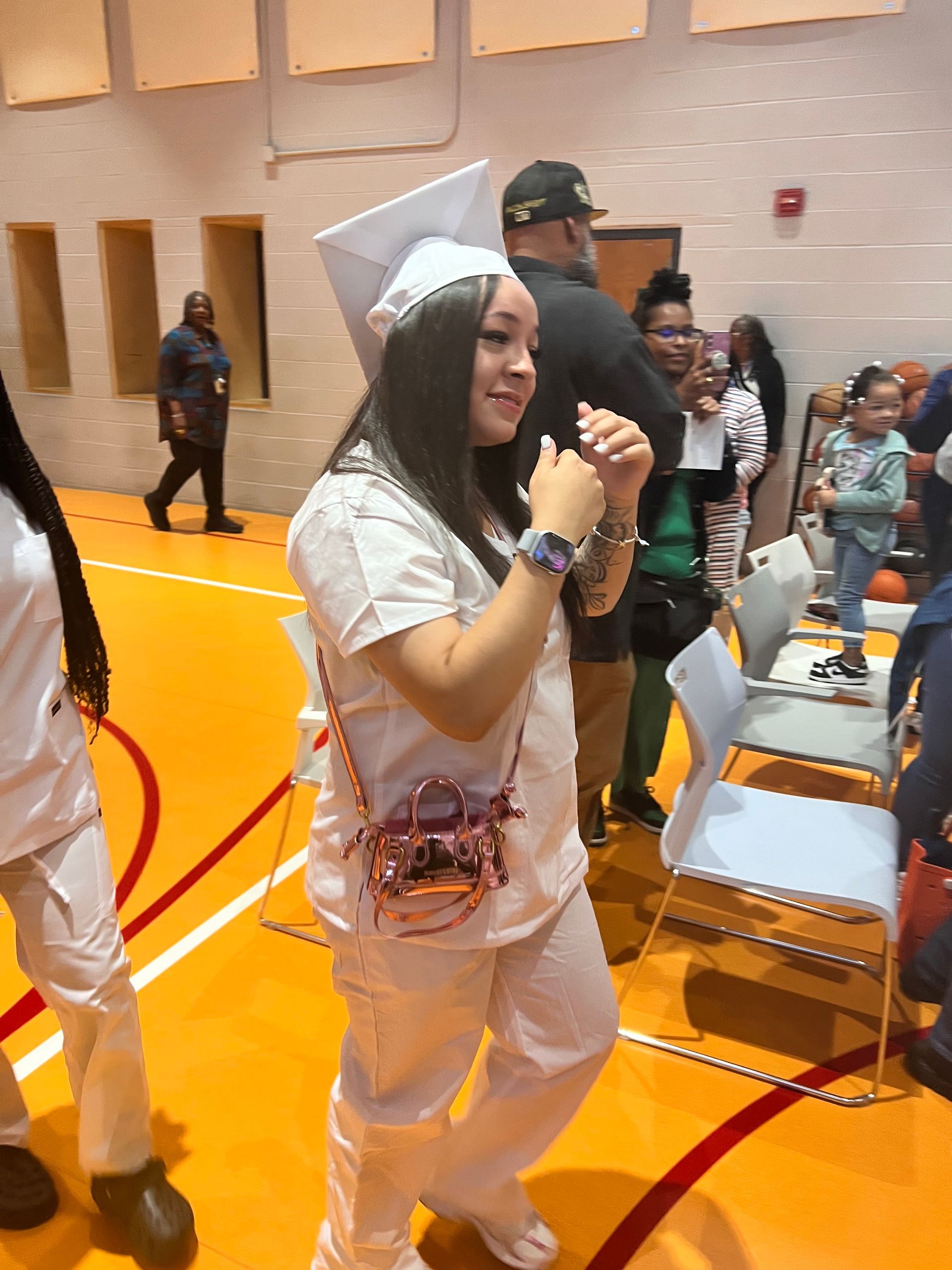 Person in white and silver outfit walking in a gymnasium, holding a phone, with chairs and seated people nearby.