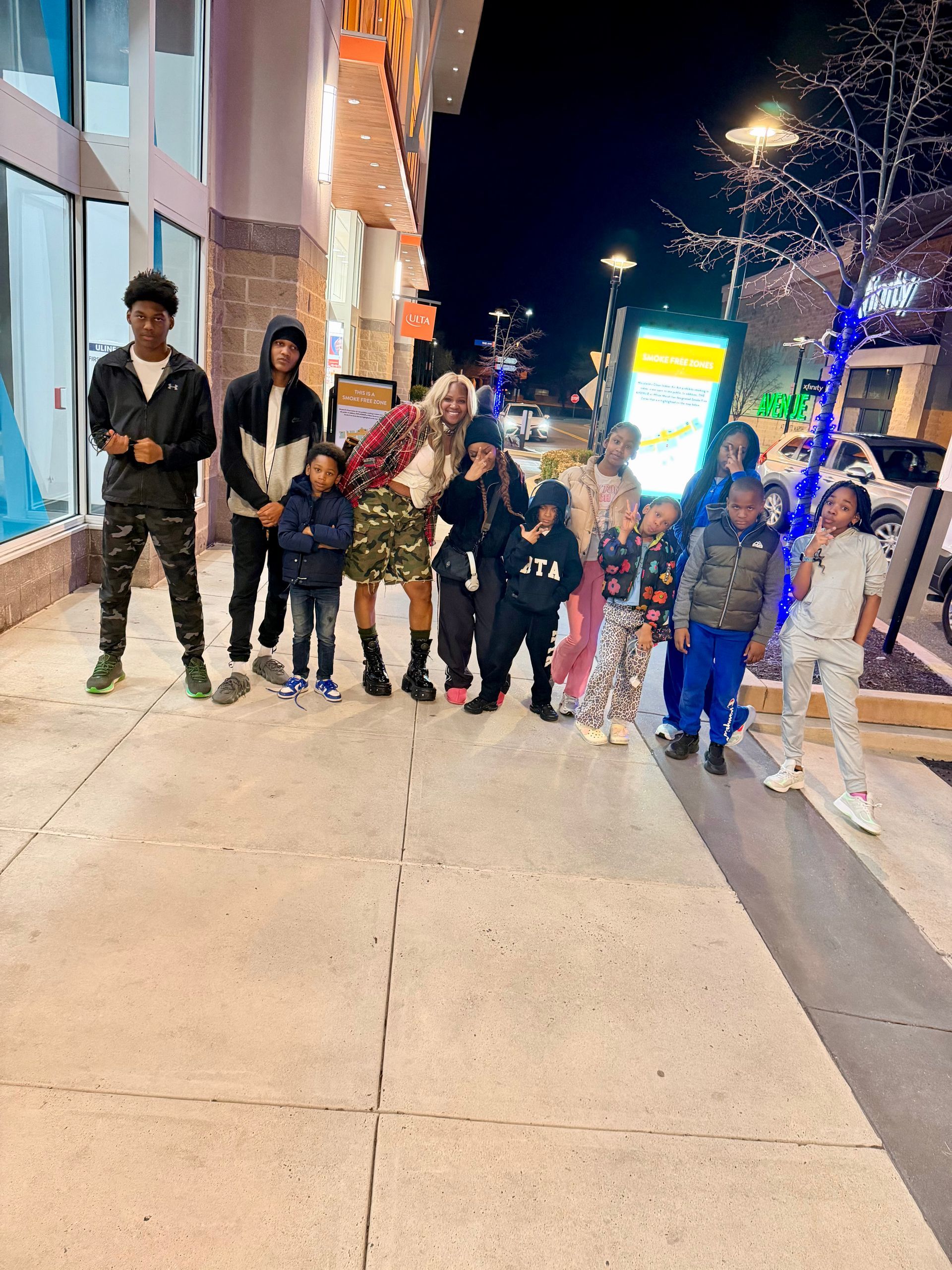 Group of people posing on a city sidewalk at night beside lit storefronts and colorful lights.