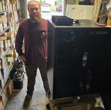 Man with beard stands next to a black server cabinet in a cluttered garage, looking at the camera.