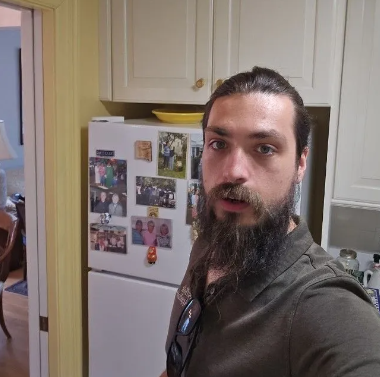 Man with beard and hair pulled back in a kitchen, standing in front of a refrigerator with photos.