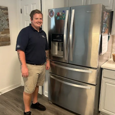 Man in a dark blue shirt and khaki shorts smiles next to a stainless steel refrigerator in a kitchen.