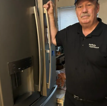 Man holding a refrigerator door handle next to a dark fridge. He wears a black shirt that says 