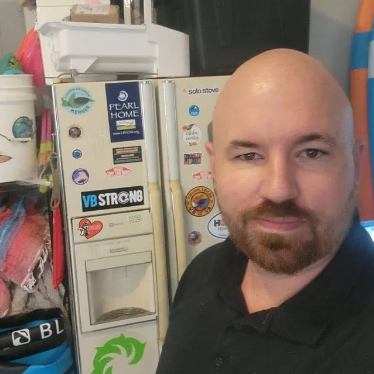Man with beard, bald head, stands beside a refrigerator covered in stickers.