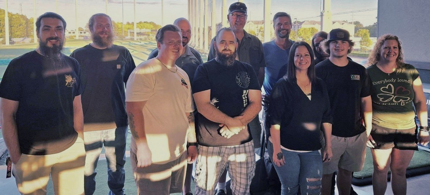 Group of people smiling, posing outdoors at a golf range. Mostly men, wearing casual clothes. Bright, sunny day.