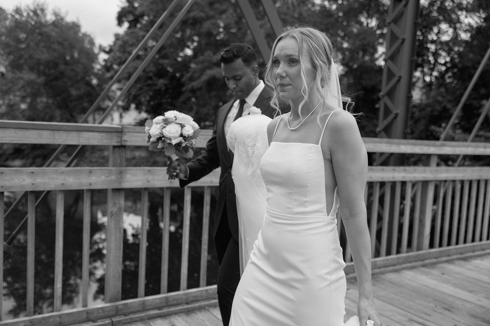 A black and white photo of a bride and groom walking across a bridge.