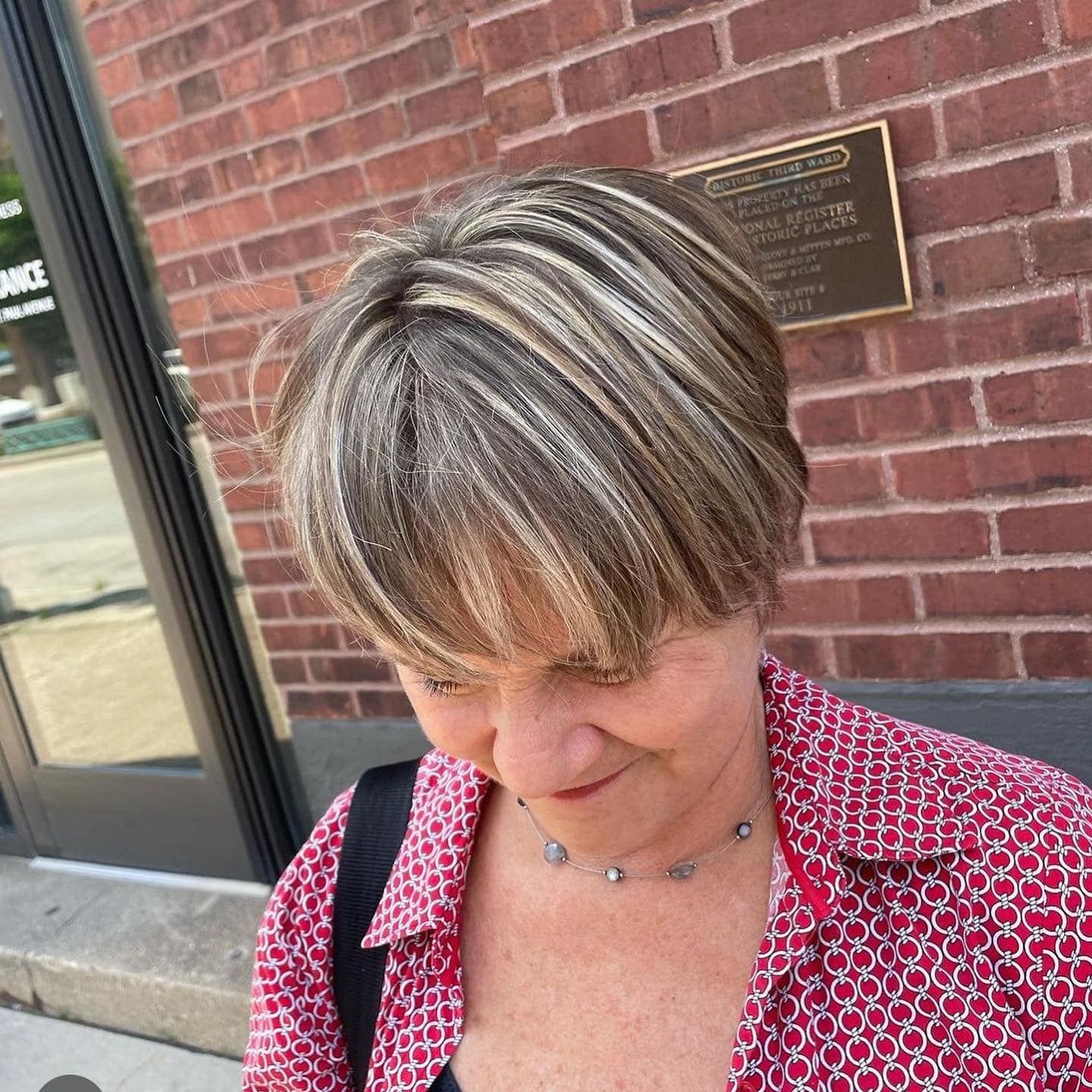 A woman wearing a red shirt and a necklace is standing in front of a brick building.