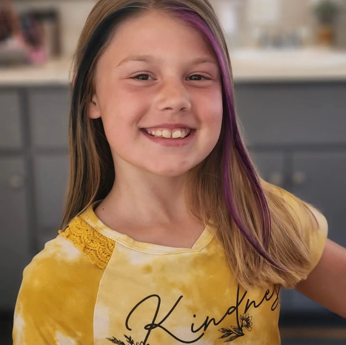A young girl with purple hair is wearing a yellow shirt that says kindness