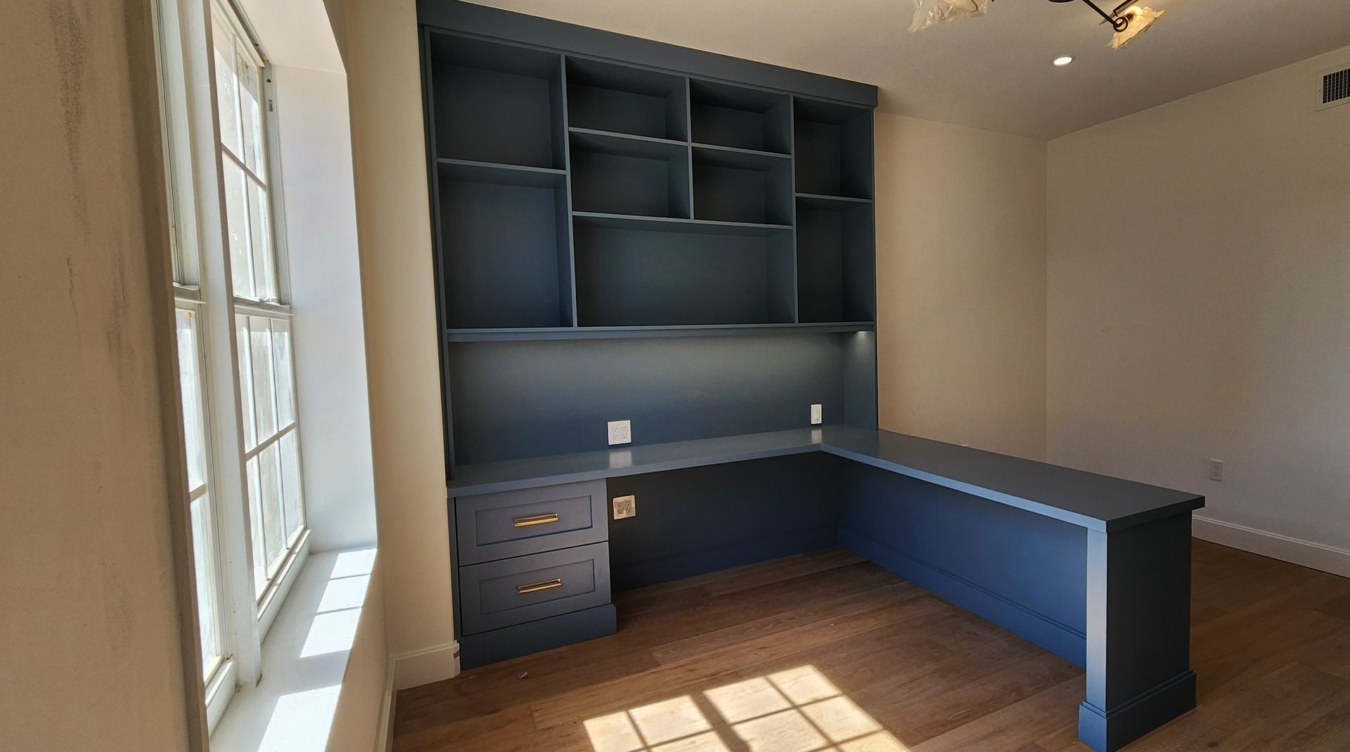 Empty home office with built-in blue desk, shelves, drawers, and sunlight from a window