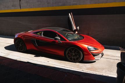 A sleek, metallic orange McLaren sports car parked on a concrete surface against a dark, two-toned wall.