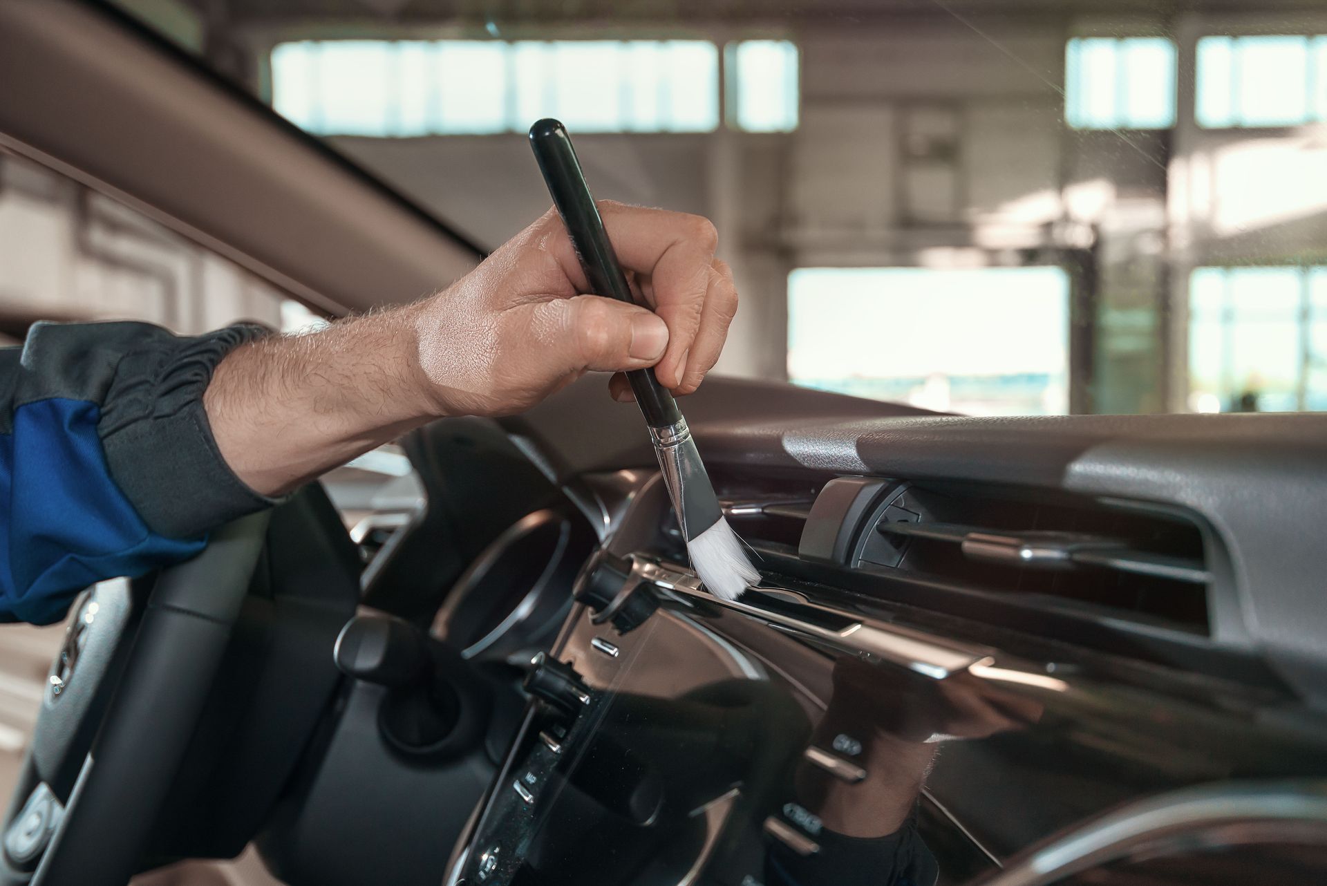 Hand using a brush to dust a car's dashboard in a well-lit garage.