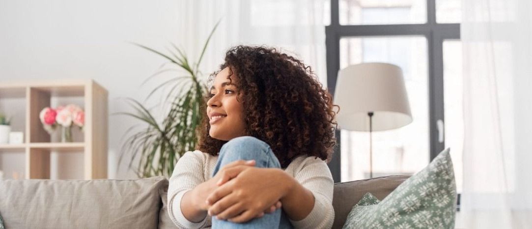 A woman is sitting on a couch hugging her knees and smiling.