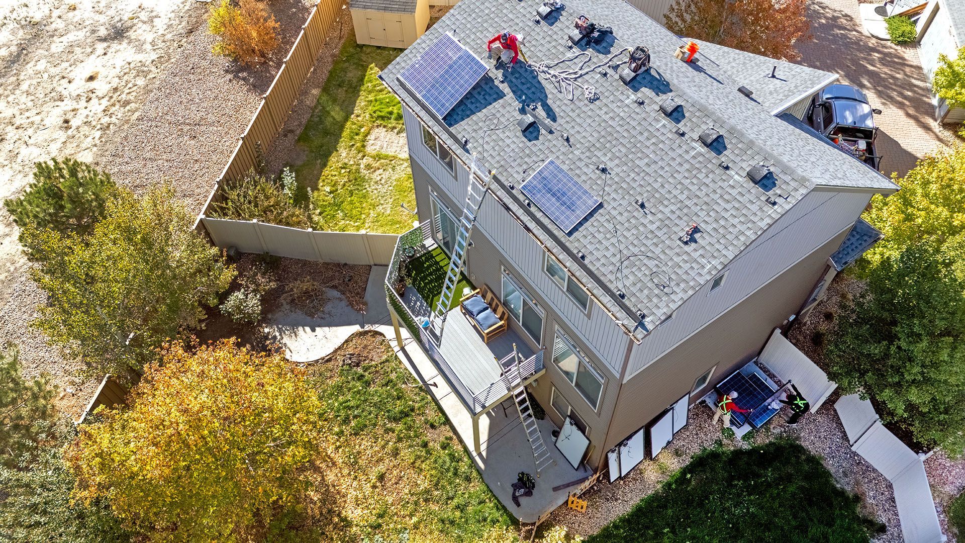 Aerial view of a two-story house with solar panels on the roof, surrounded by a yard with trees and a fence.