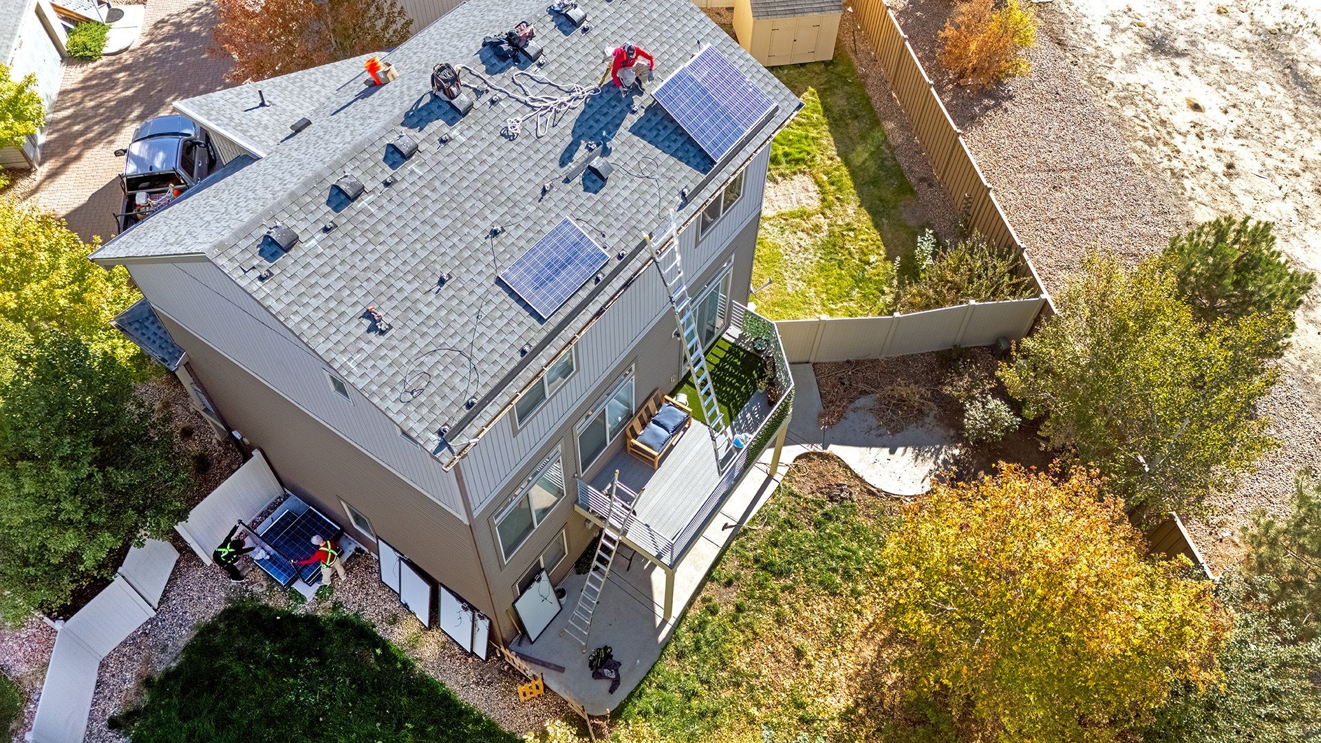High-angle view of a two-story house with solar panels on the roof and a backyard with trees.