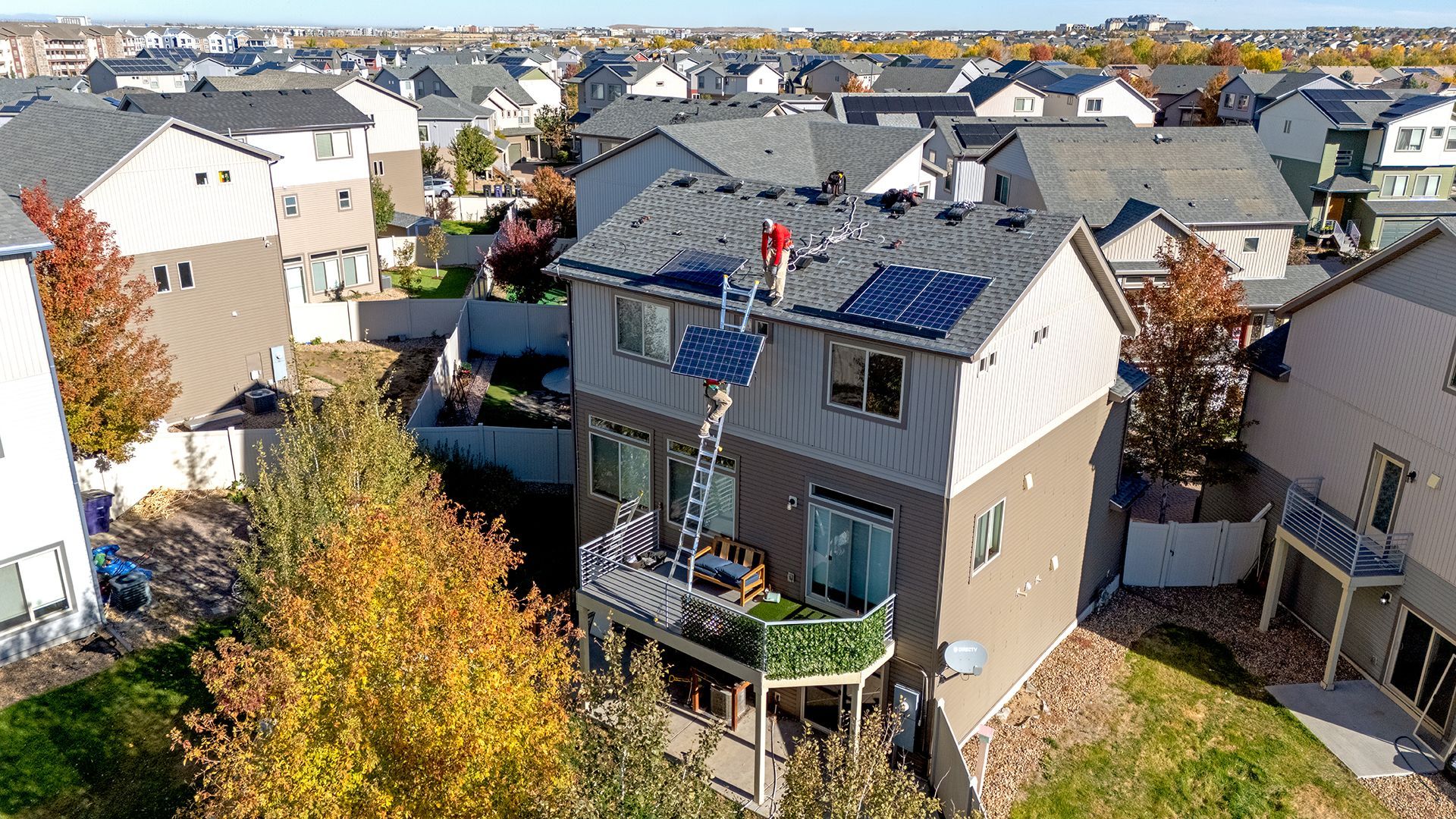 Solar panel installation on a two-story house roof, worker on a ladder, suburban setting.