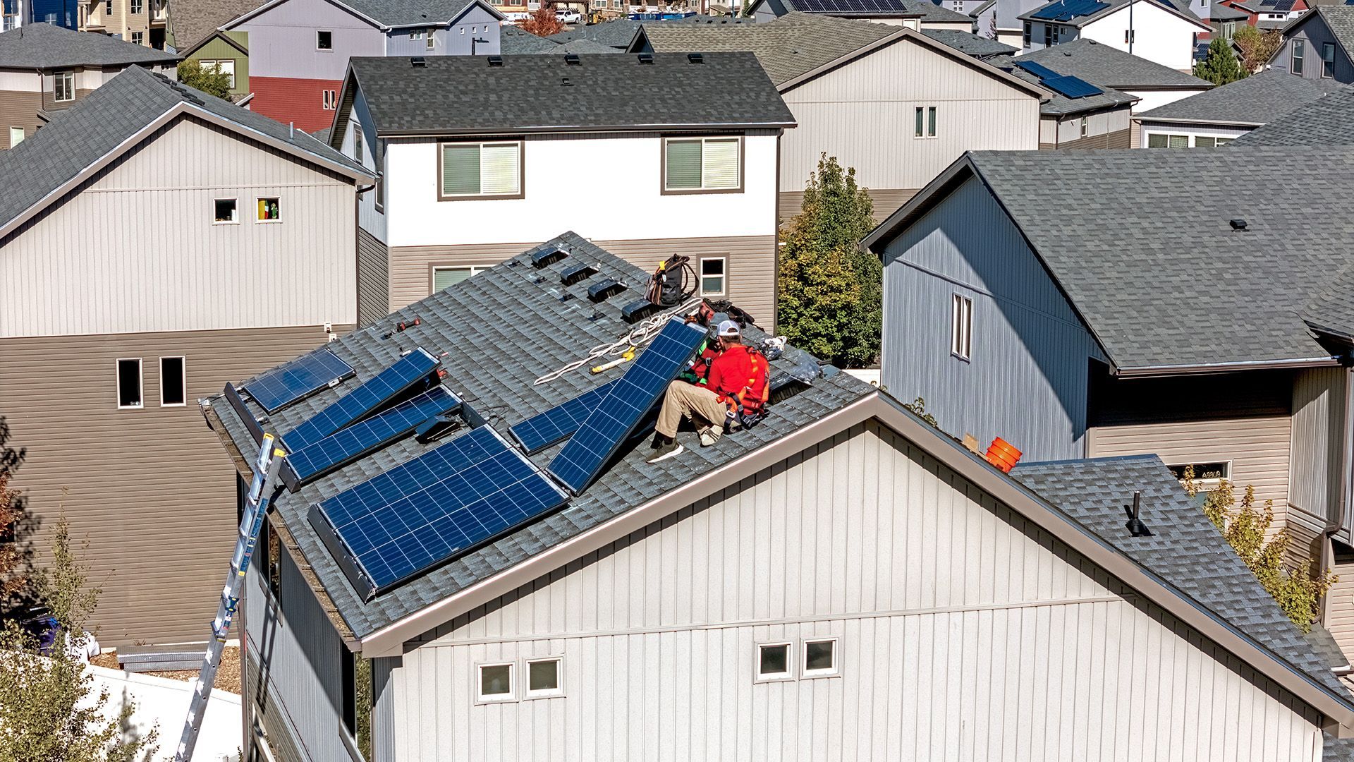 Workers installing solar panels on a residential rooftop, surrounded by similar houses.