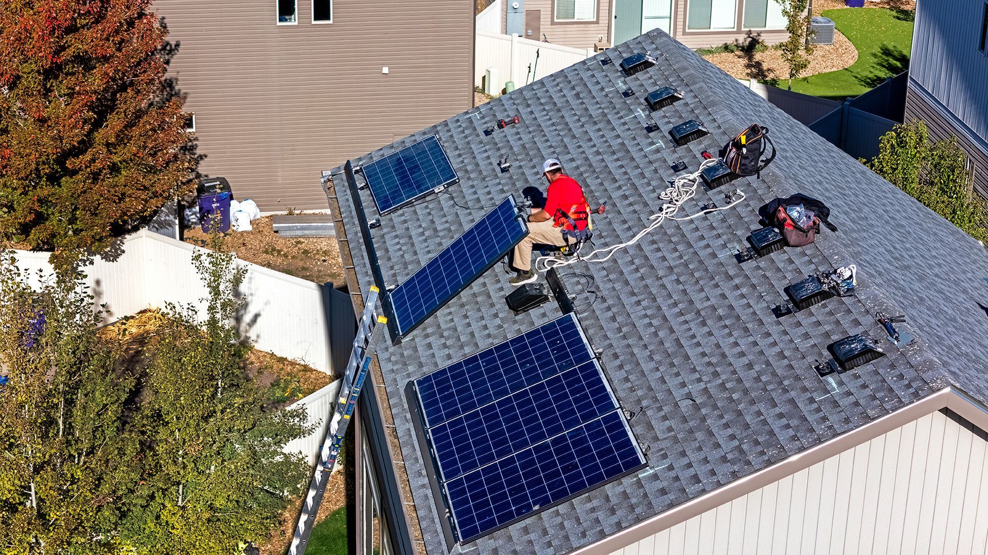 Workers installing solar panels on a residential rooftop. Blue panels, gray roof, man in red shirt.