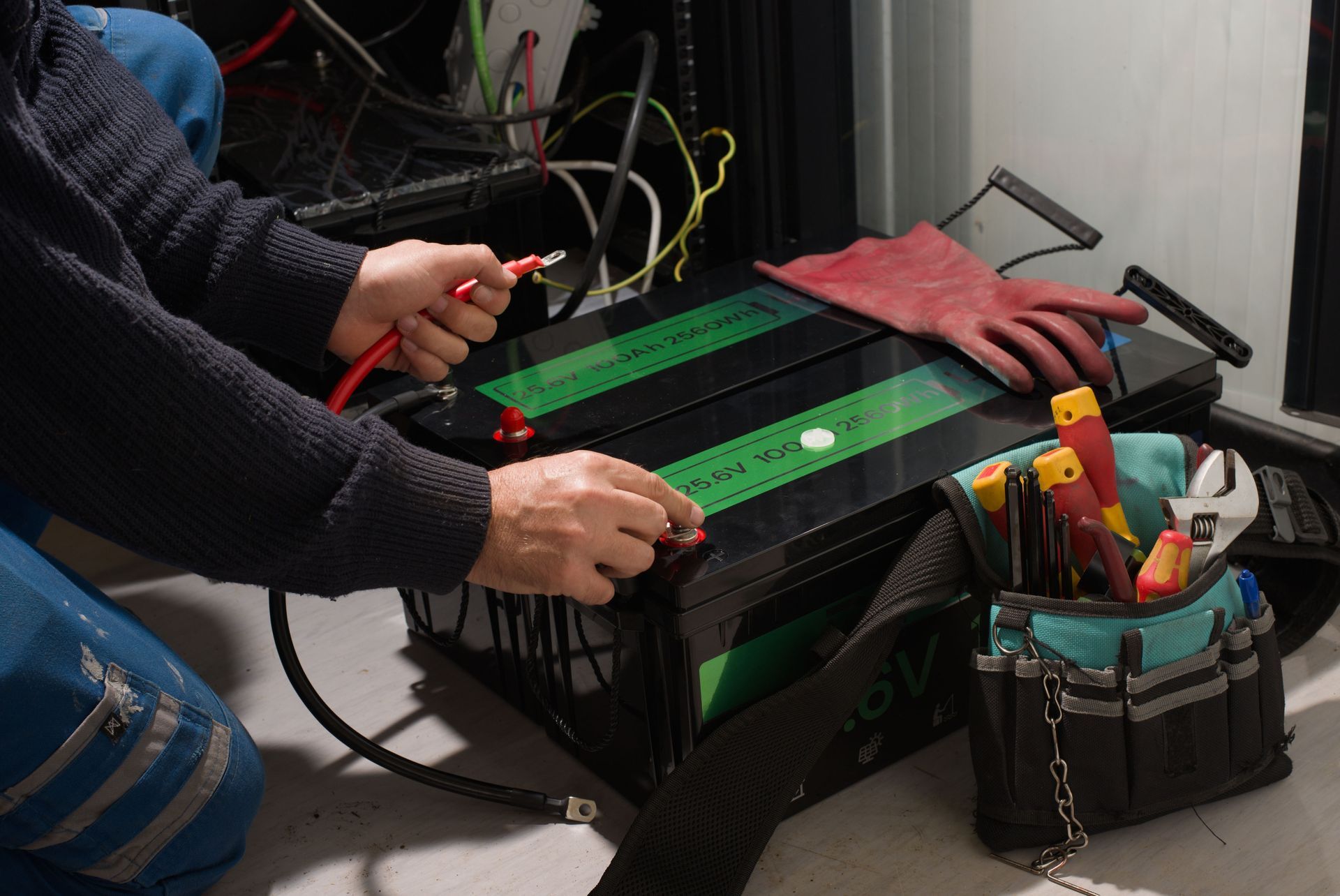 Person connecting a red cable to a battery. Tools and a glove are in a nearby bag.