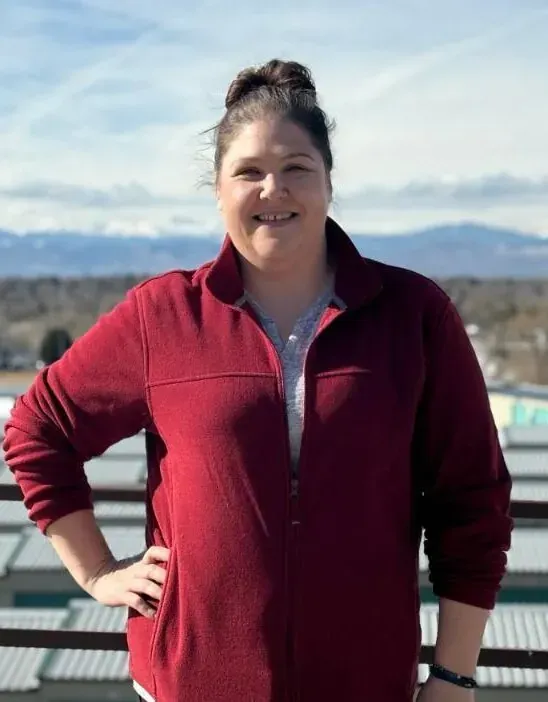Woman in red fleece jacket, smiling, hand on hip, against a backdrop of mountains and a blue sky.