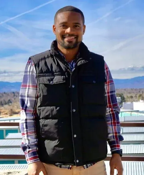 Man wearing a black vest and plaid shirt smiles outdoors.