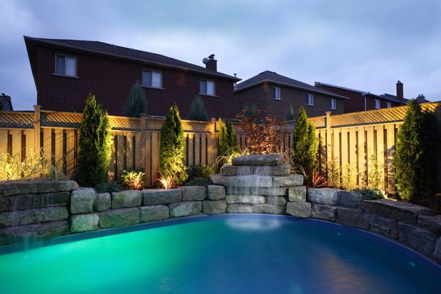 Nighttime backyard pool with waterfall, lit by spotlights. Stone wall, wooden fence, and tall trees.