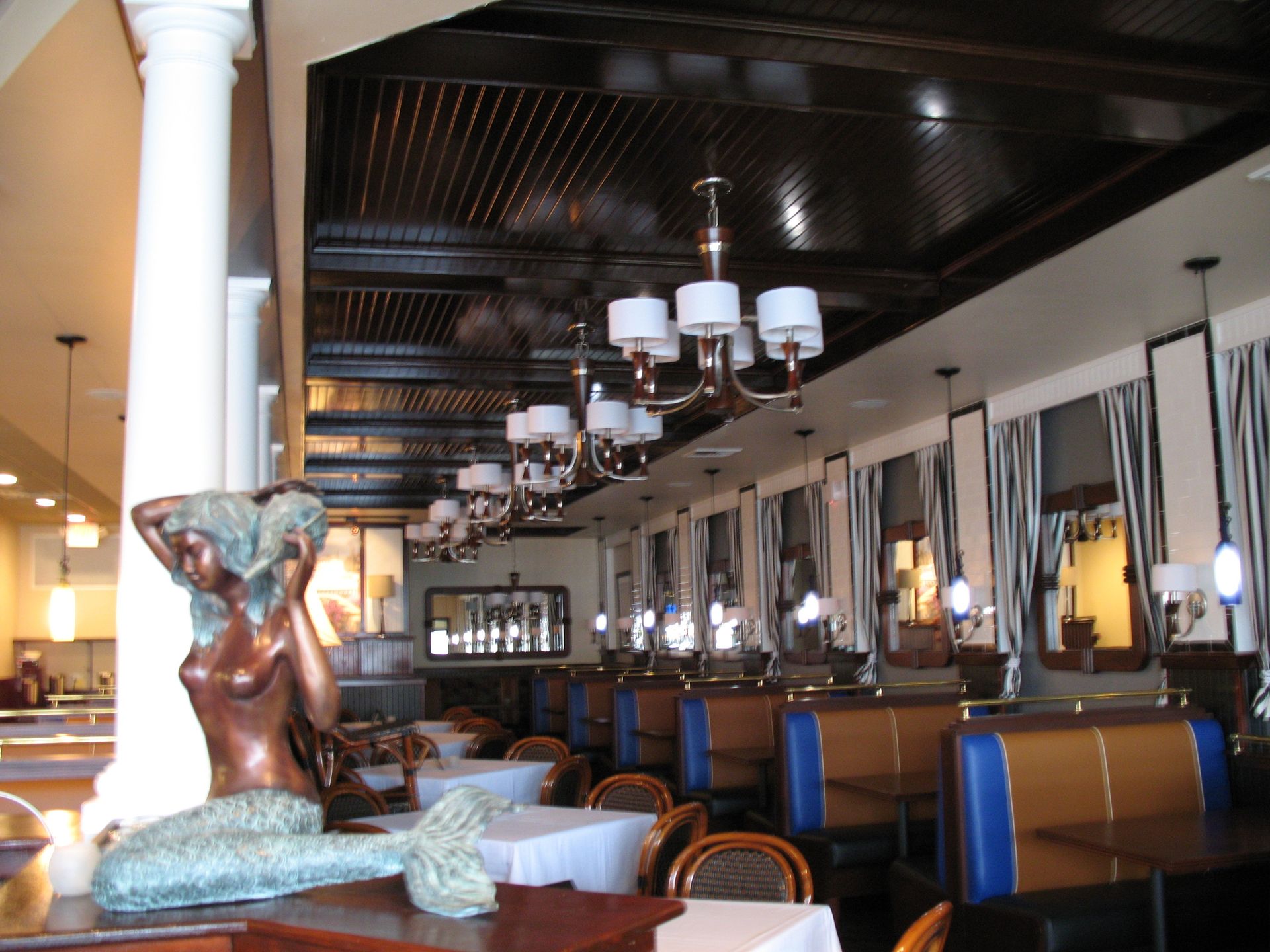 Restaurant interior with mermaid statue, booths, white tablecloths, chandeliers, and dark wood ceiling.