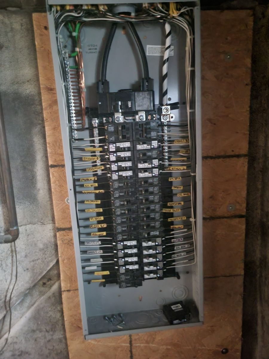 Electrical panel with numerous circuit breakers, wires, and busbars. Metal enclosure mounted on a wooden surface.