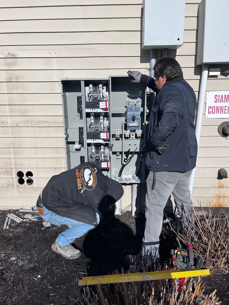 Two electricians working on an electrical panel box mounted on a building exterior.
