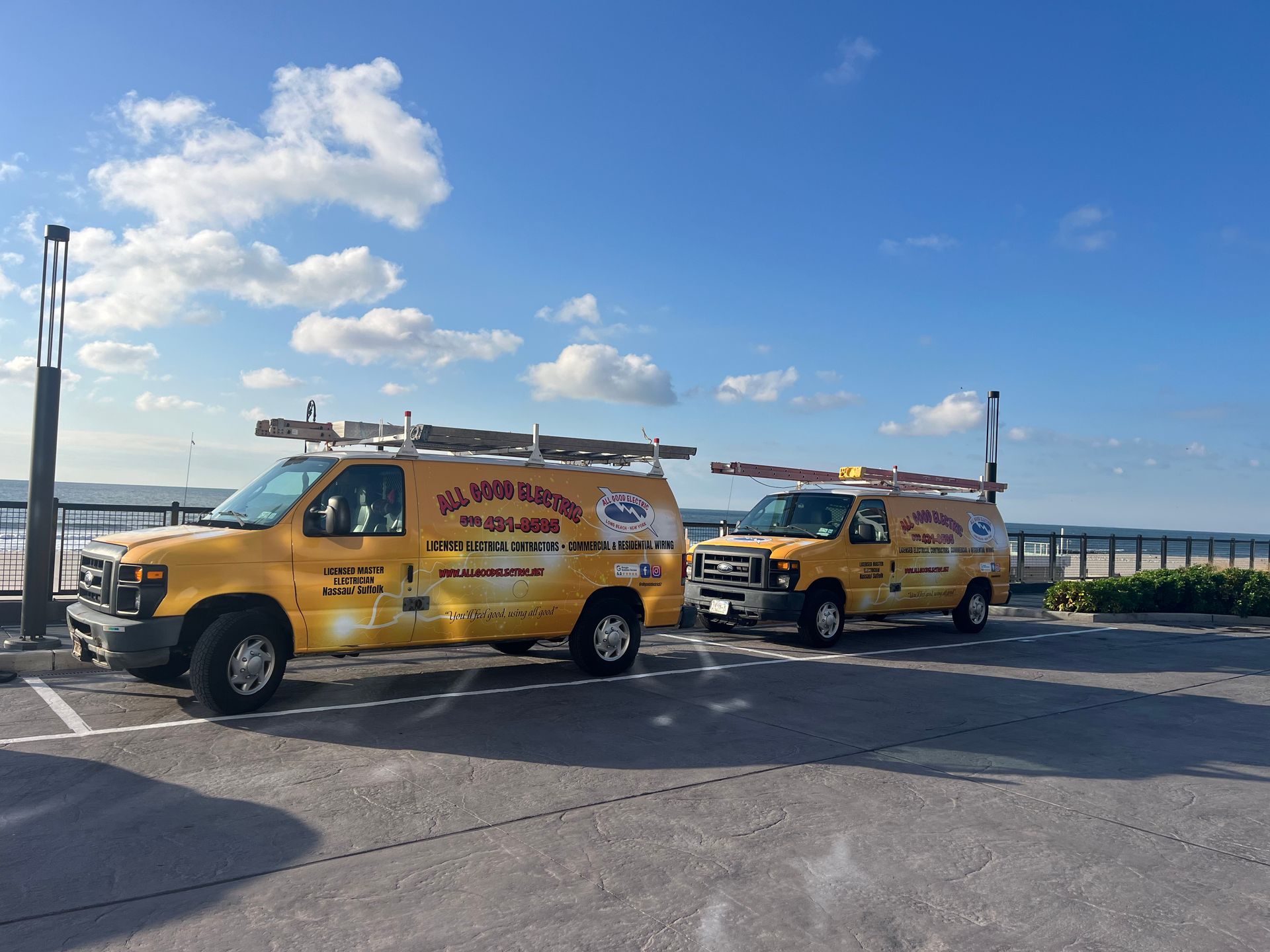 Two yellow service vans parked, both with ladders, near water under a blue sky.