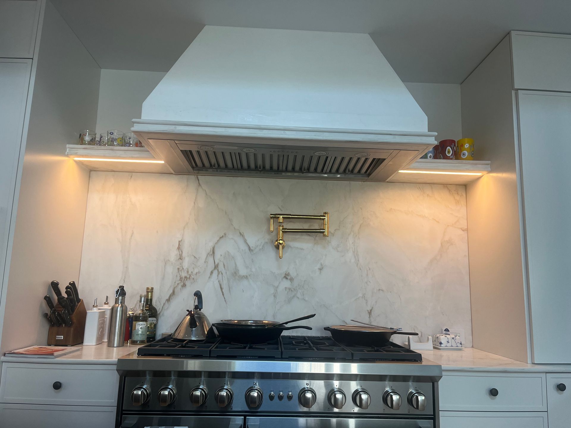 White kitchen with a stove, hood, and marble backsplash. Shelves and under-cabinet lighting.