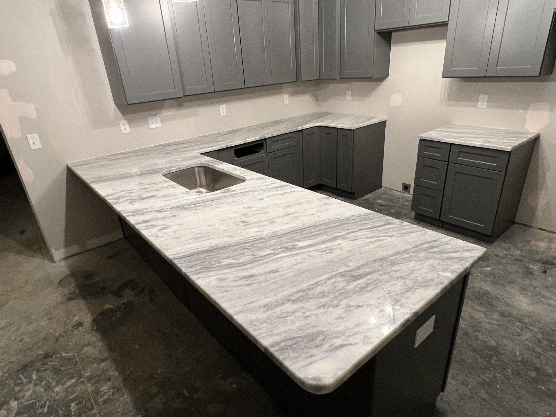 Gray kitchen cabinets and island with light-colored countertop, unfinished walls and floor.