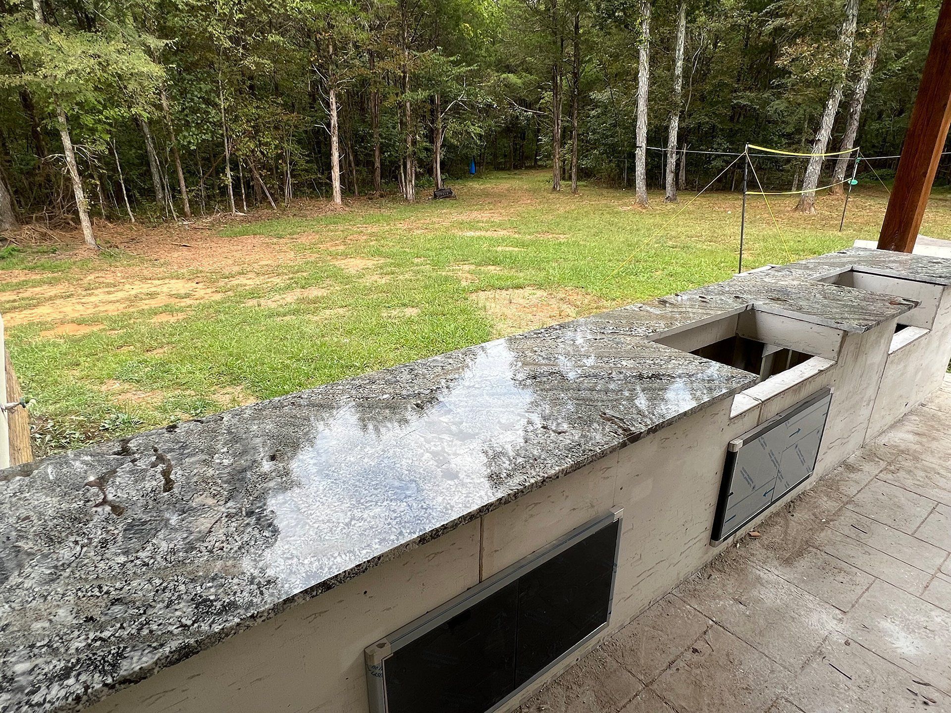 Outdoor granite countertop with a built-in sink and storage, overlooking a grassy yard and trees.