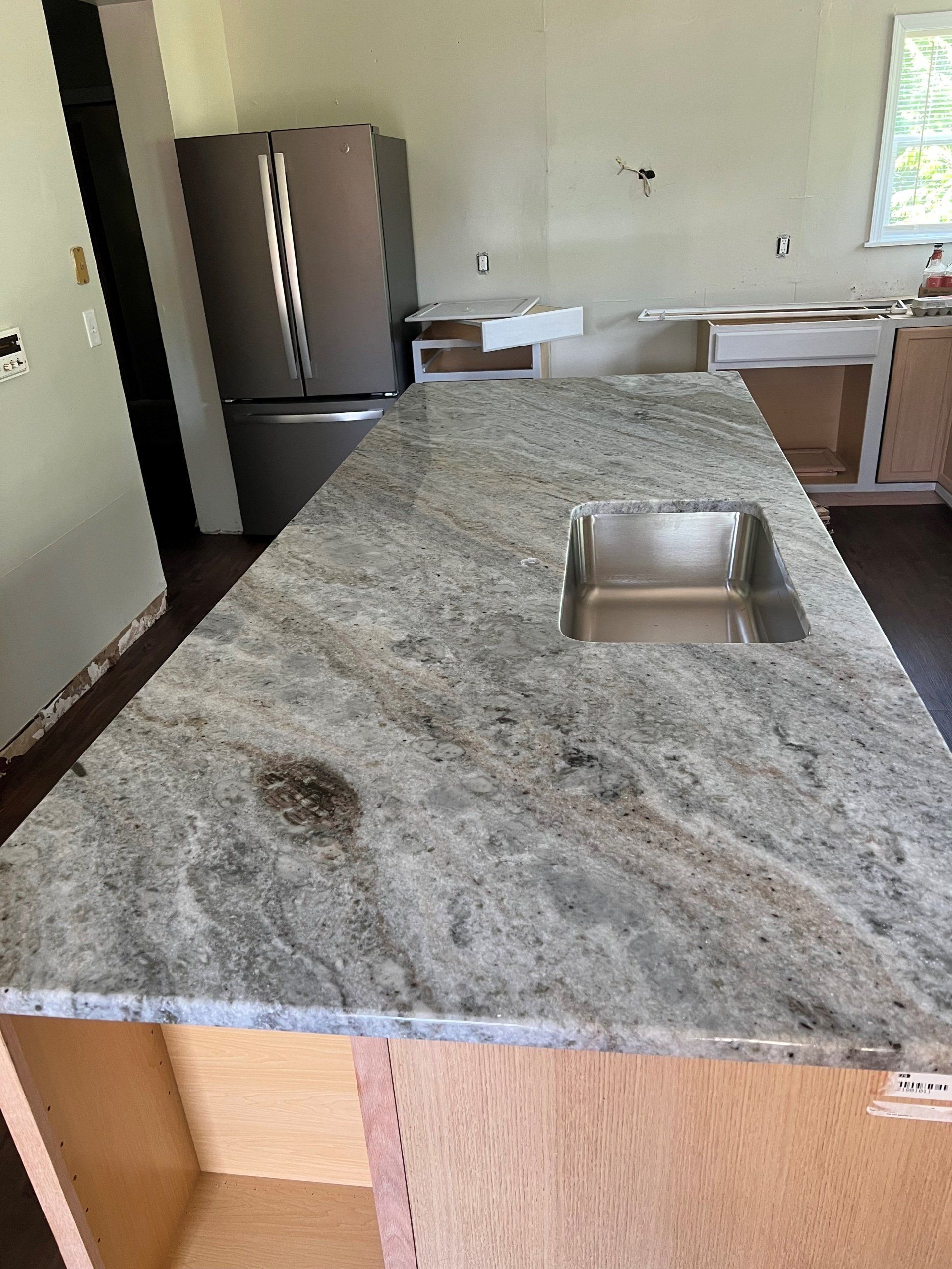 Kitchen island with granite countertop and stainless steel sink, unfinished cabinets, refrigerator in background.