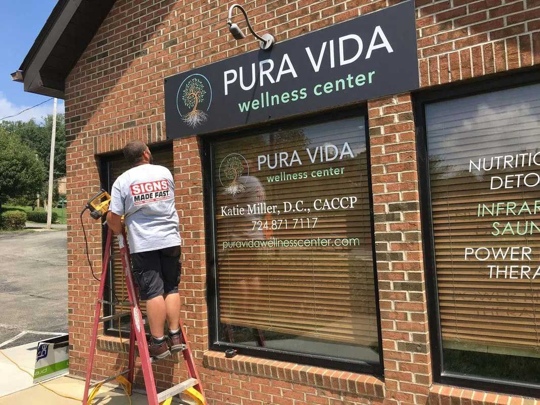 A person on a ladder installs a window sign for Pura Vida Wellness Center, a brick building with a black sign.