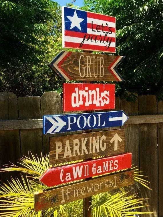 Wooden directional signs for a party in a backyard. Signs point to food, drinks, pool, parking, lawn games, and fireworks.