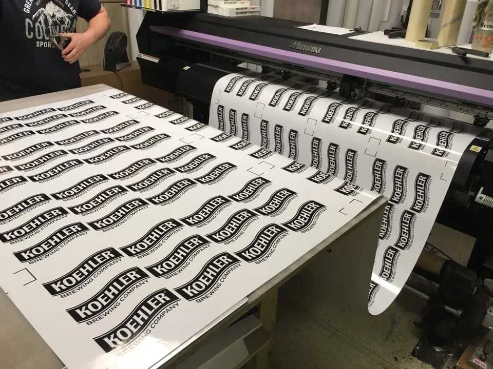 A roll-to-roll printer producing adhesive labels in black and white with a logo, in a print shop.
