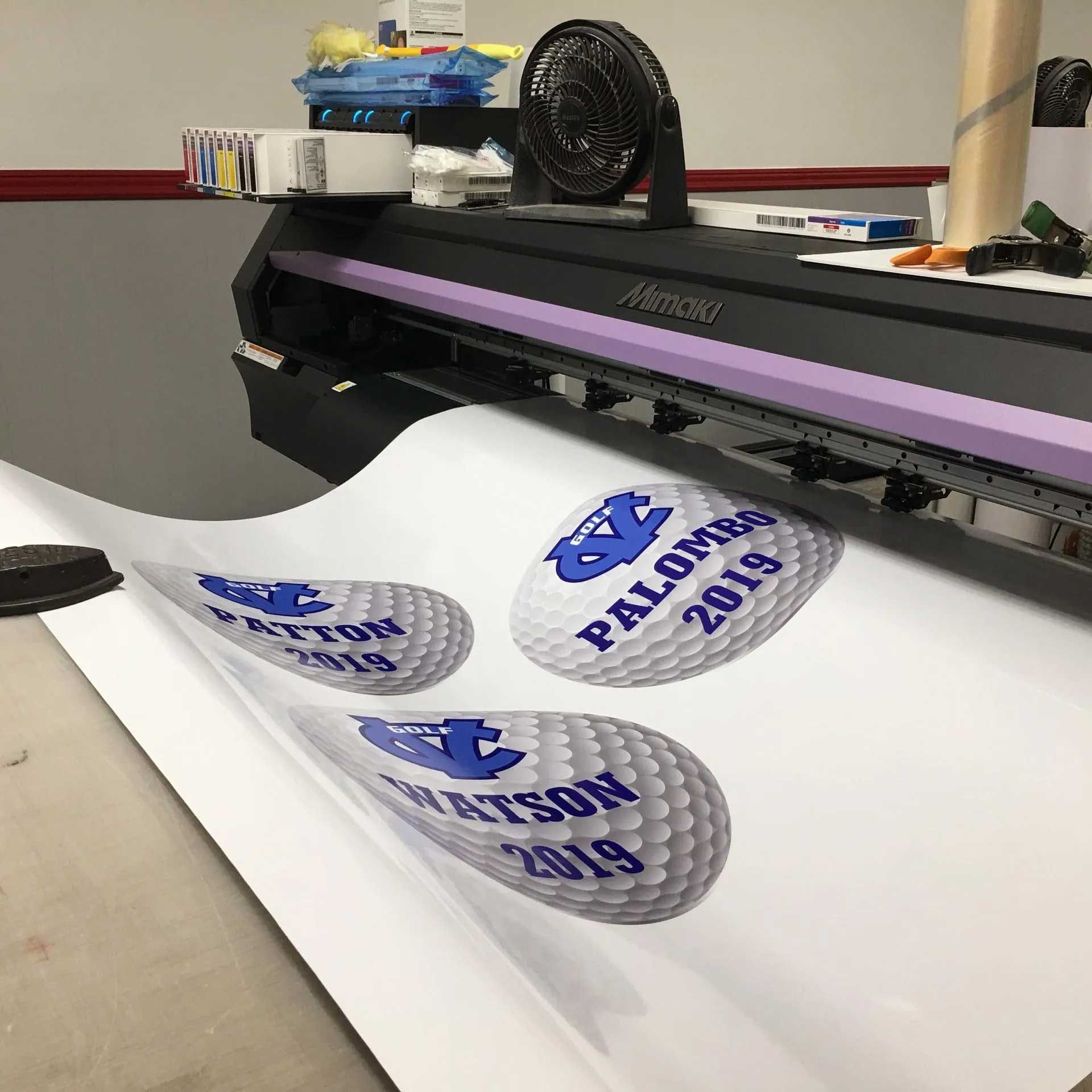 A printer producing white vinyl decals with blue text and a golf ball design.