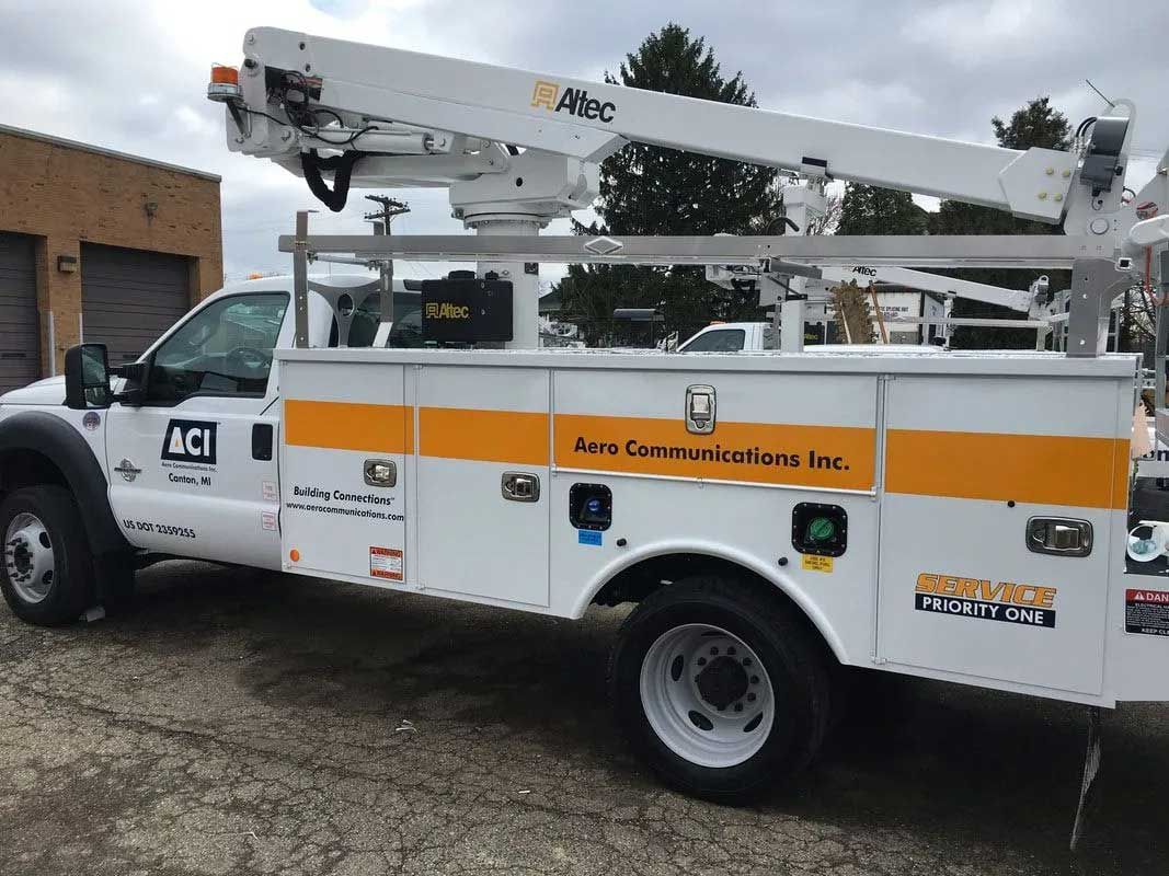 White utility truck with an aerial lift, parked outdoors. The truck has orange stripes and storage compartments.